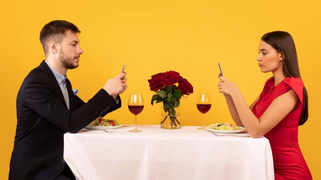 A man and woman sit at a dinner table with wine, both looking at smartphones.