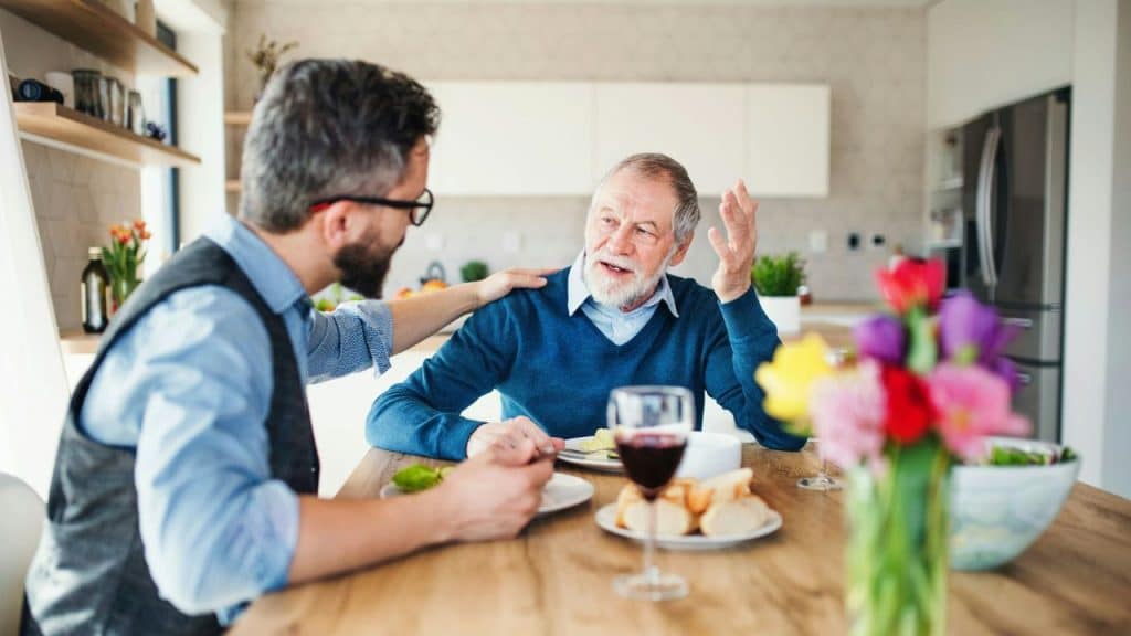 Two men sitting at a dining table talking, with one man gesturing with his hand.