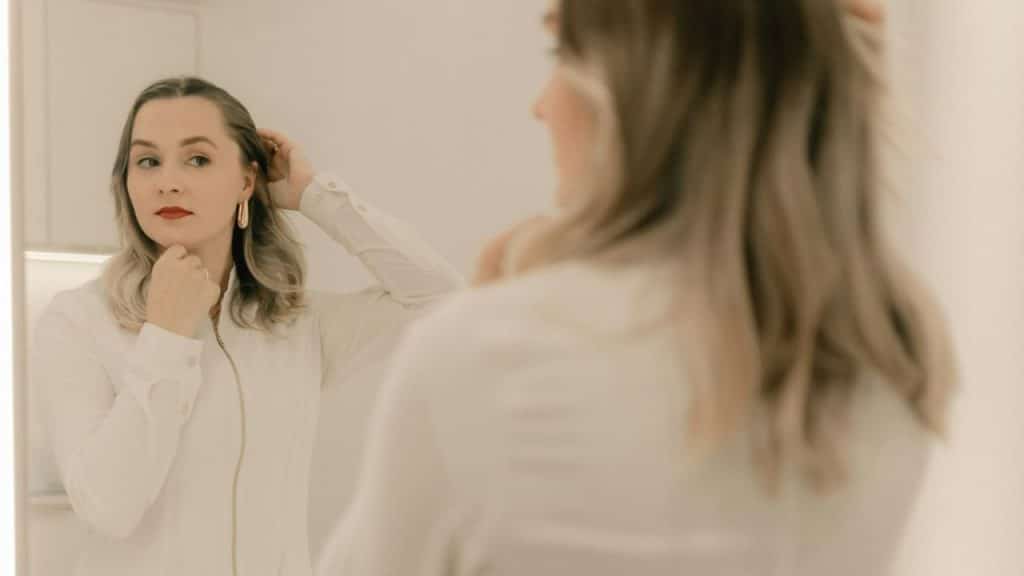 A woman in a white shirt looks at her reflection in a mirror while touching hair.