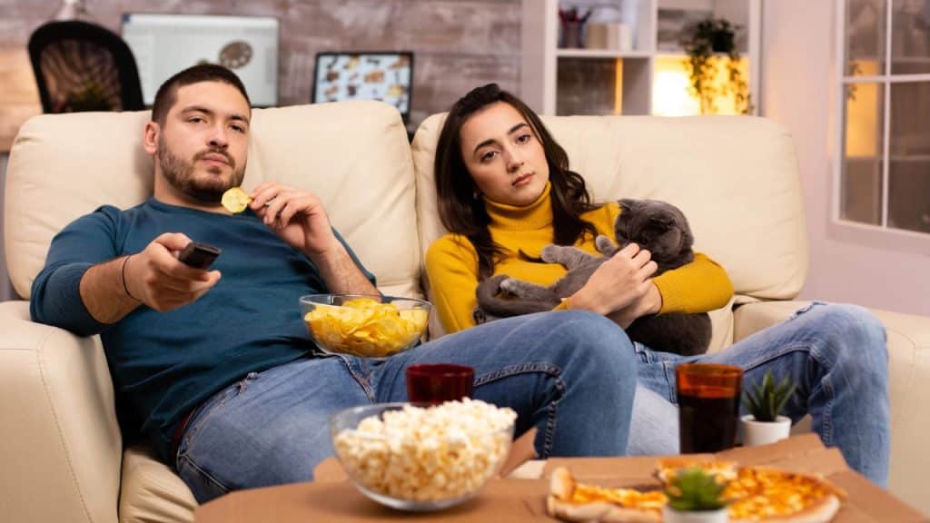A man and woman sit on a couch with a cat, popcorn, and pizza.
