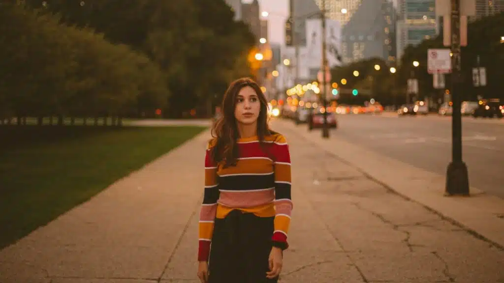 A woman in a striped sweater walks on a sidewalk with city lights behind her.
