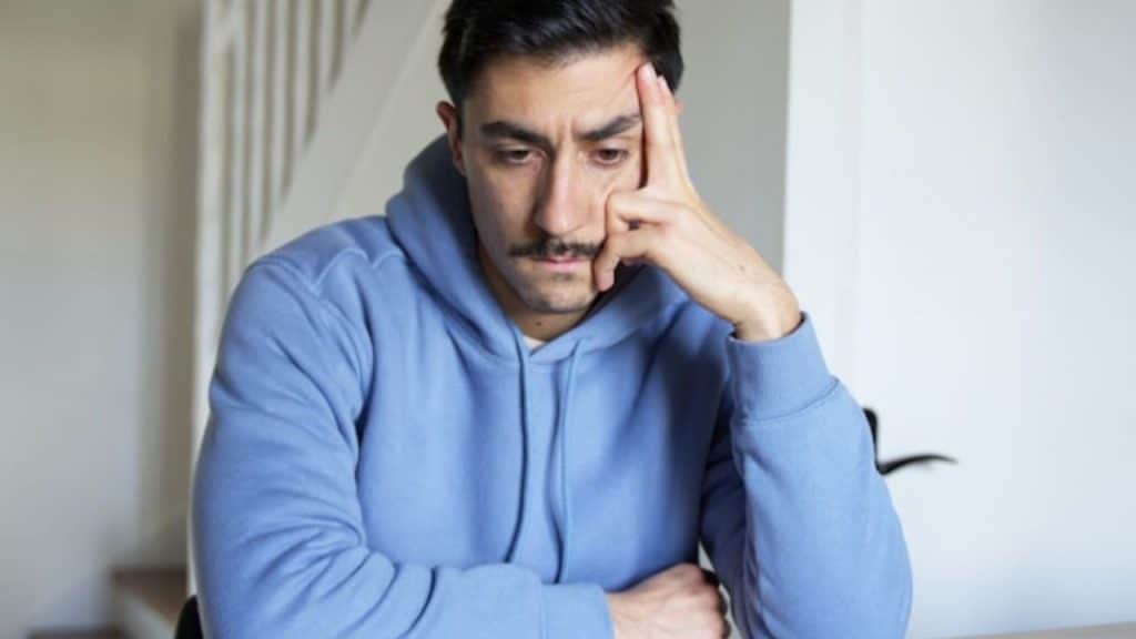 A man with a mustache wearing a blue hoodie resting his head on his hand.
