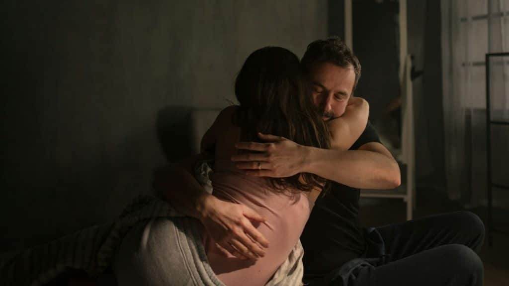 A man and woman embrace in a dimly lit room while sitting on a bed.