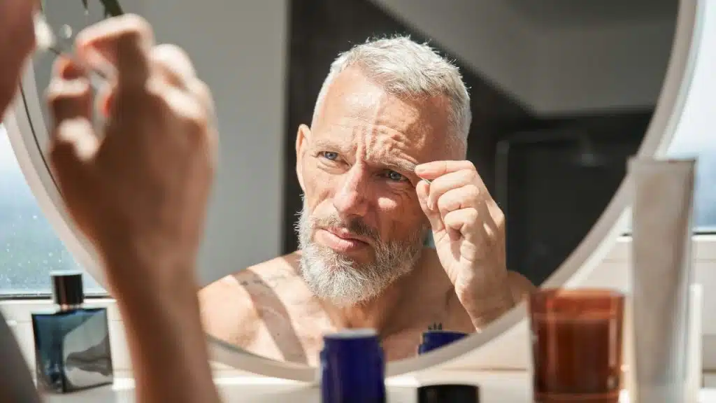 A man with a grey beard uses tweezers on his eyebrow while looking in a mirror.