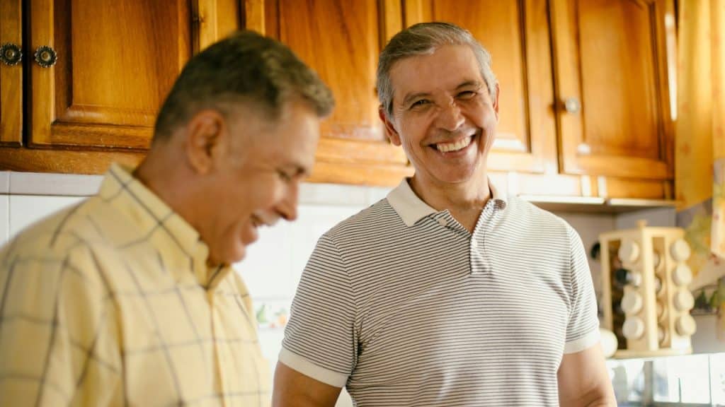 Two men stand in a kitchen with wooden cabinets; one is smiling broadly at the camera.