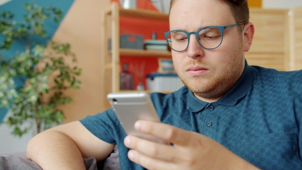 A man with blue glasses and a polo shirt looks down at a silver smartphone.