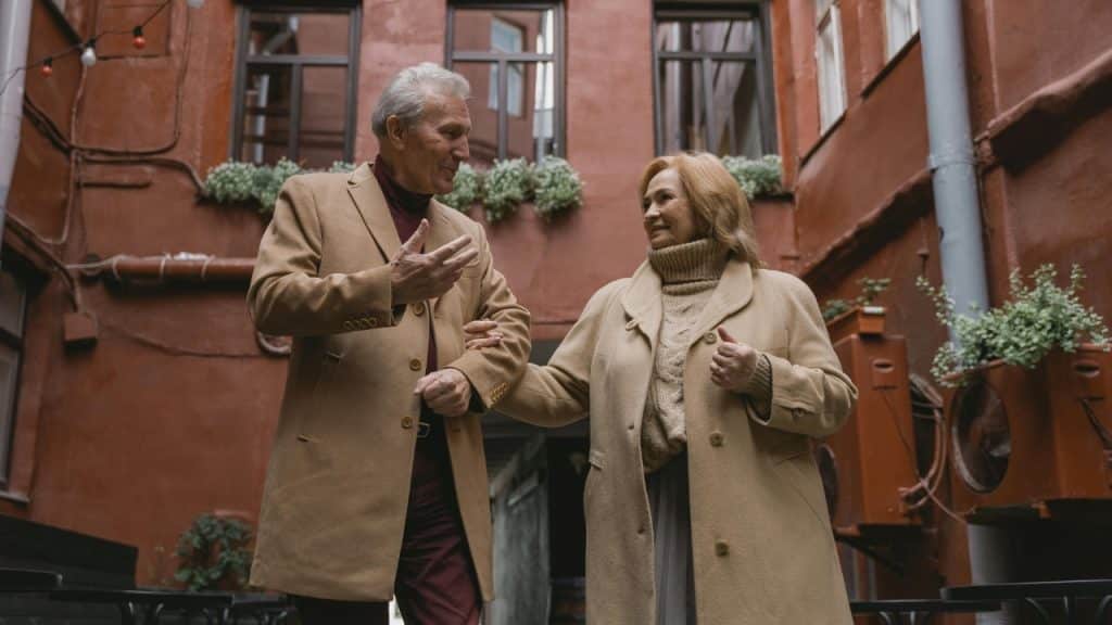 An older man and woman in tan coats walk together through a brick-walled courtyard.