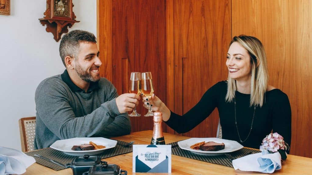 A man and woman sit at a dinner table, smiling while clinking champagne glasses together.