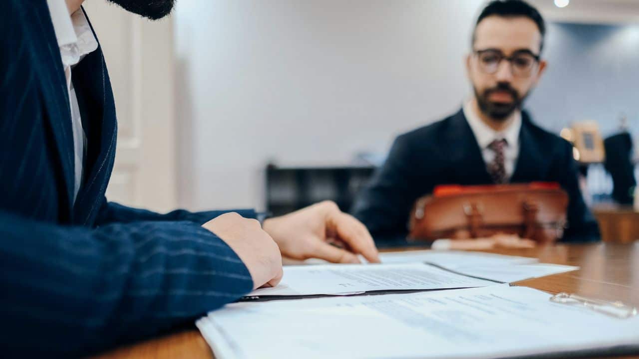 Two people listen to a man in a suit explain information from a white document.