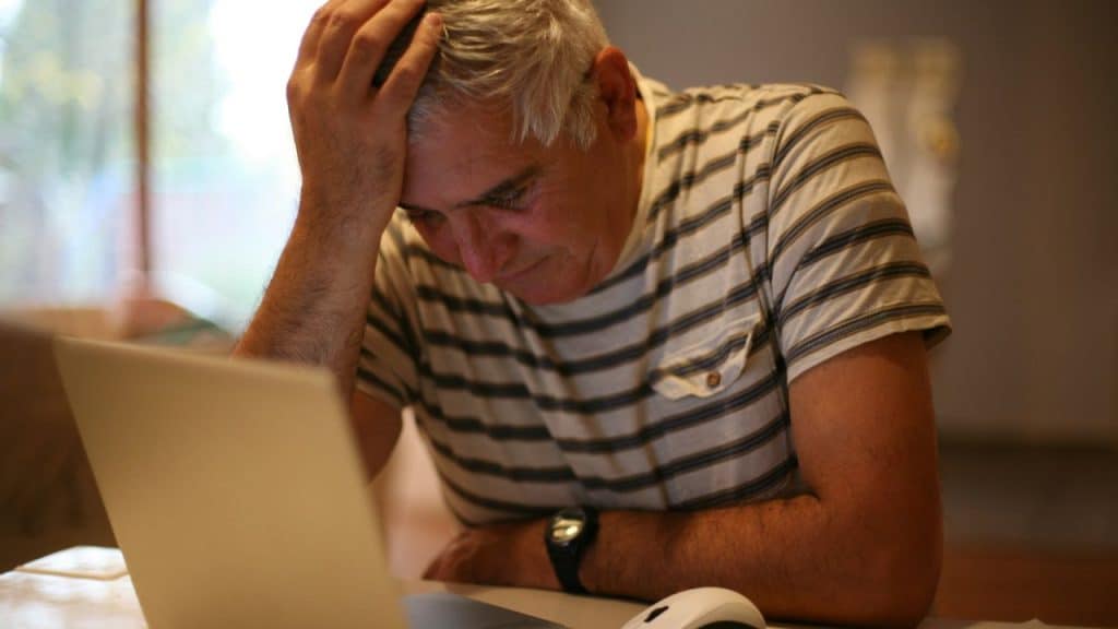 A man with gray hair sits at a desk, resting his forehead on his hand.