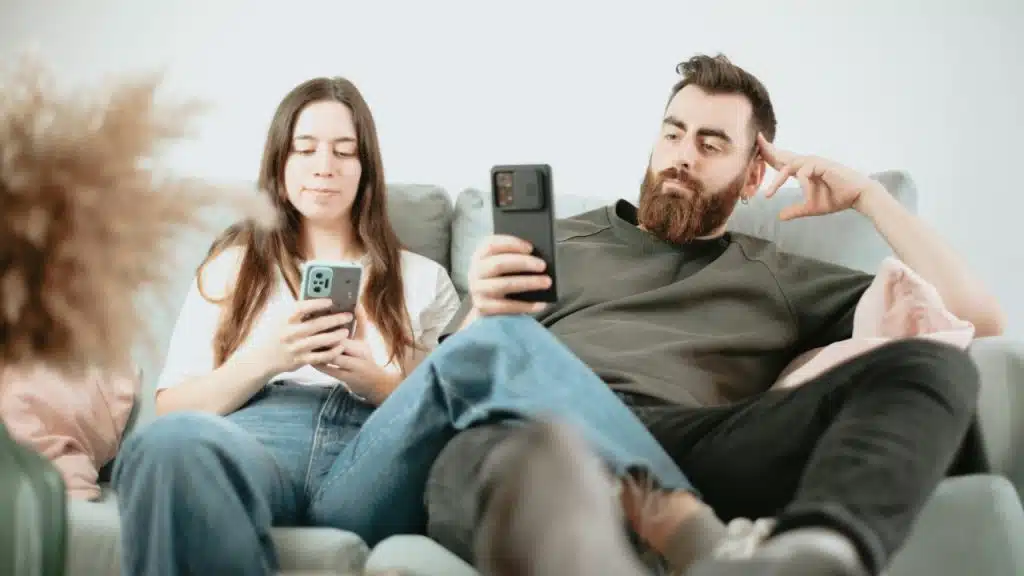 A man and woman sit on a couch, both looking down at their individual smartphones.