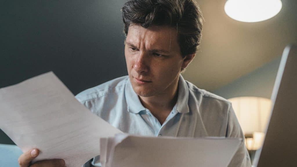 A man with dark hair looks down intently while holding and reviewing several white papers.