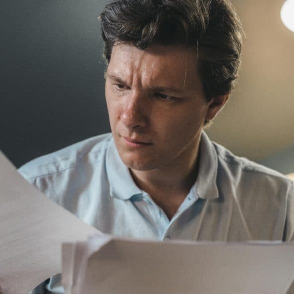 A man with dark hair looks down intently while holding and reviewing several white papers.