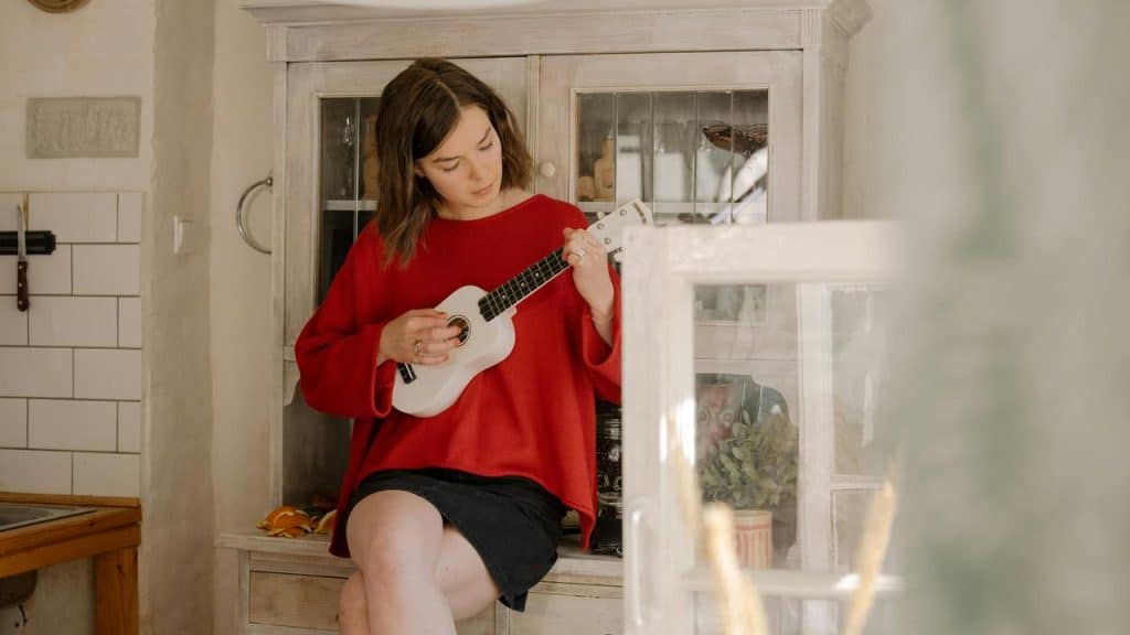 A woman in a red sweater playing a small ukulele while sitting indoors.