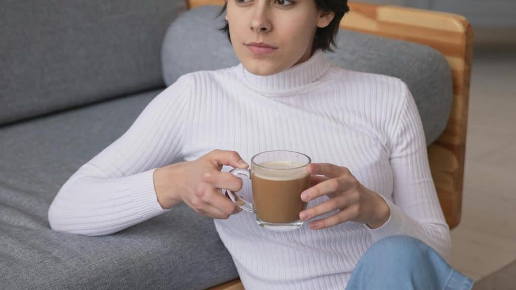A woman in a white sweater sitting on a couch holding a cup of coffee.