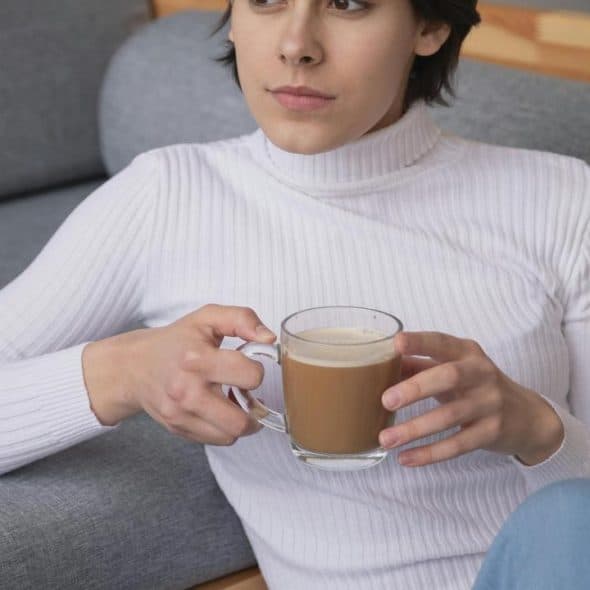 A woman in a white sweater sitting on a couch holding a cup of coffee.