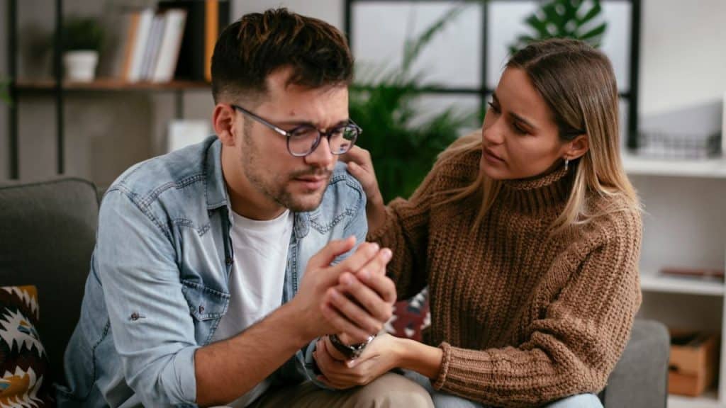 A woman rests her hand on the shoulder of a man who is looking down thoughtfully.