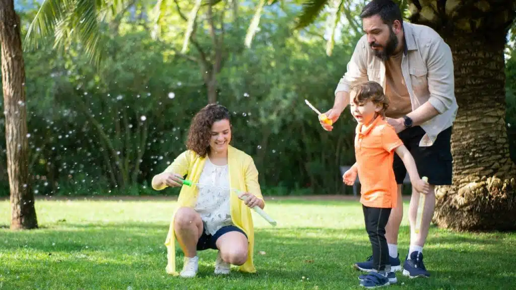 A woman and man play with a young boy blowing bubbles in a grassy park.