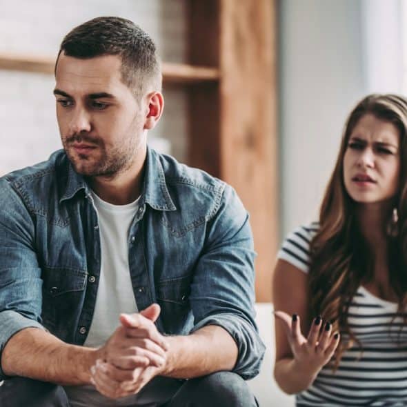 A man sitting on a couch looking away while a woman argues or gestures in frustration behind him.