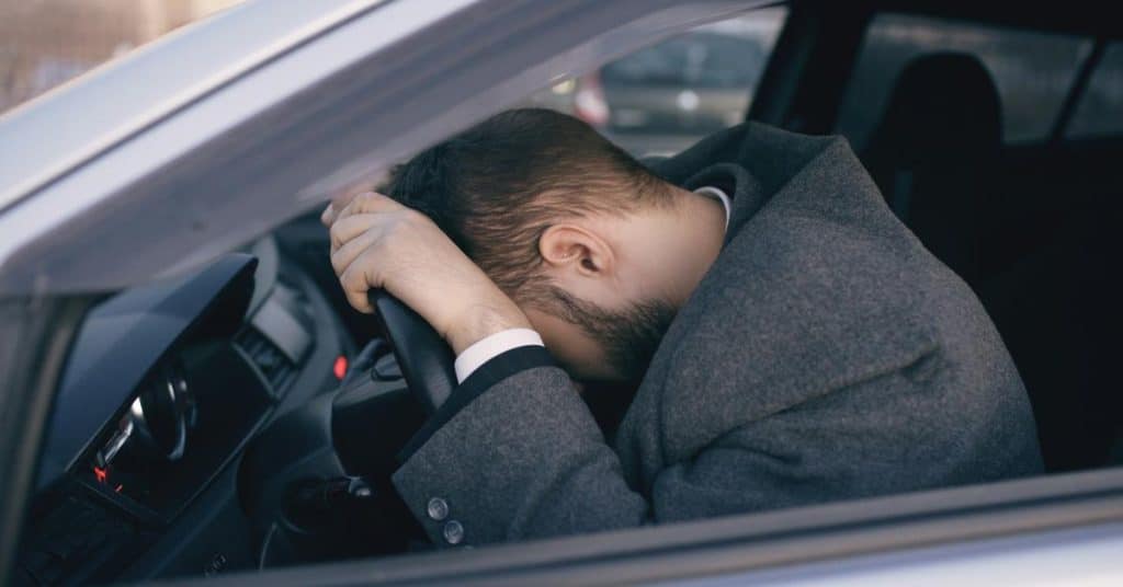 A man slumped over a car’s steering wheel, appearing exhausted.