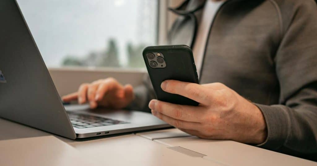 A person holds a phone while typing on a laptop.