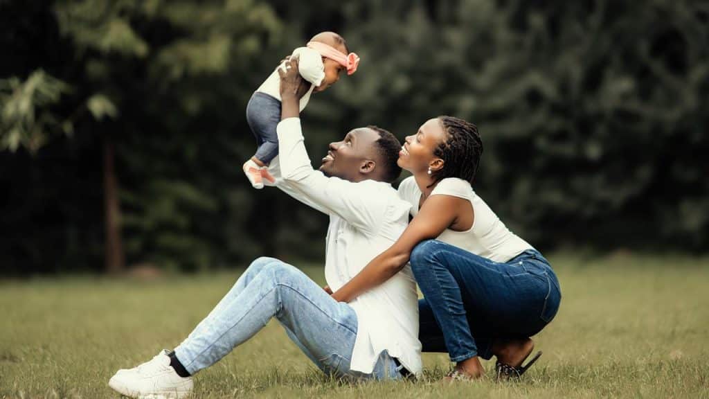 A couple smiling as the man lifts a baby into the air in a grassy field.