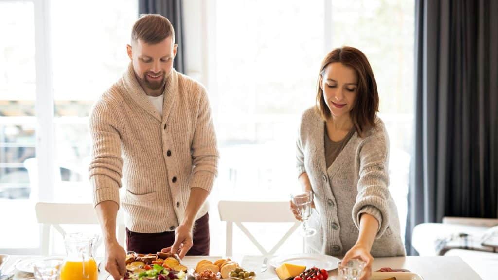 A couple setting a table with food and drinks in a bright dining room.