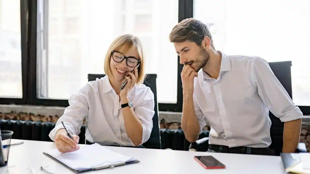 A woman on the phone writing notes while a man watches.