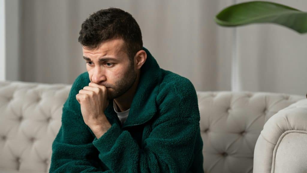 A man in a green fleece jacket sits on a white sofa looking downward pensively.