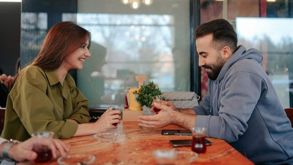 A man and woman sit at a wooden table in a cafe, talking over small glasses.