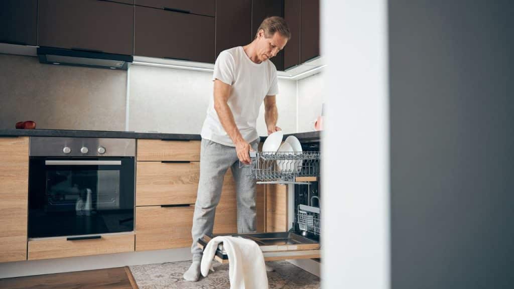 A man in a white shirt and grey pants loads white plates into a kitchen dishwasher.
