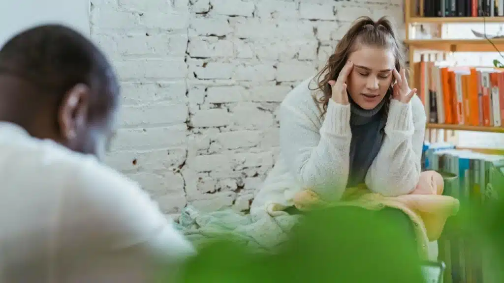 A woman in a white sweater sits cross-legged and presses her fingers to her temples.