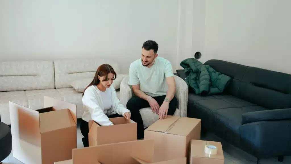 A man and woman sit on the floor and in a chair surrounded by cardboard boxes.