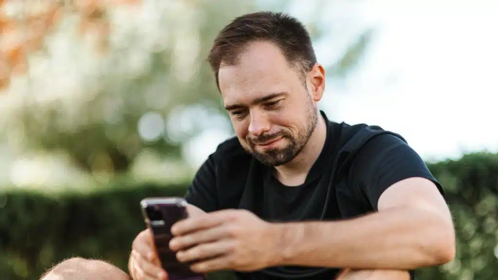 A man with a beard looks down at a smartphone while sitting outdoors during the day.