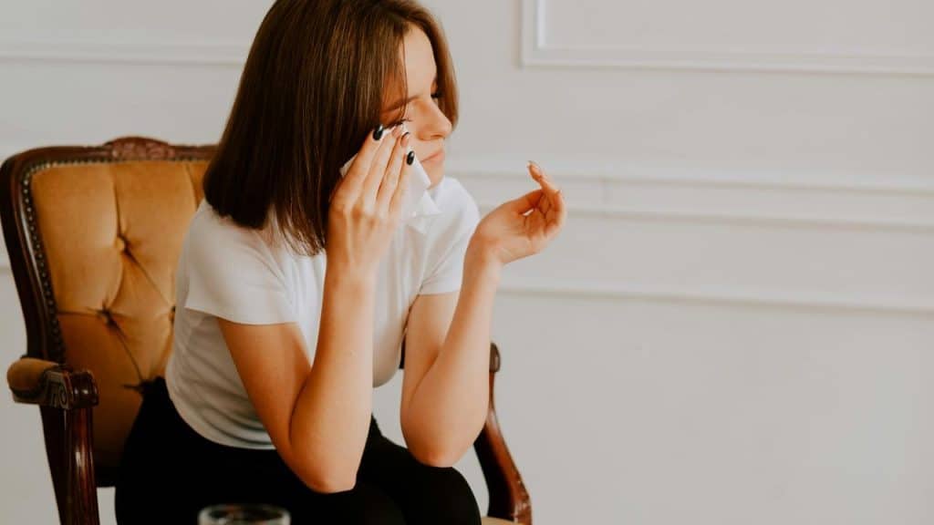 A woman sits in a chair, holding a tissue to her face with a pensive expression.