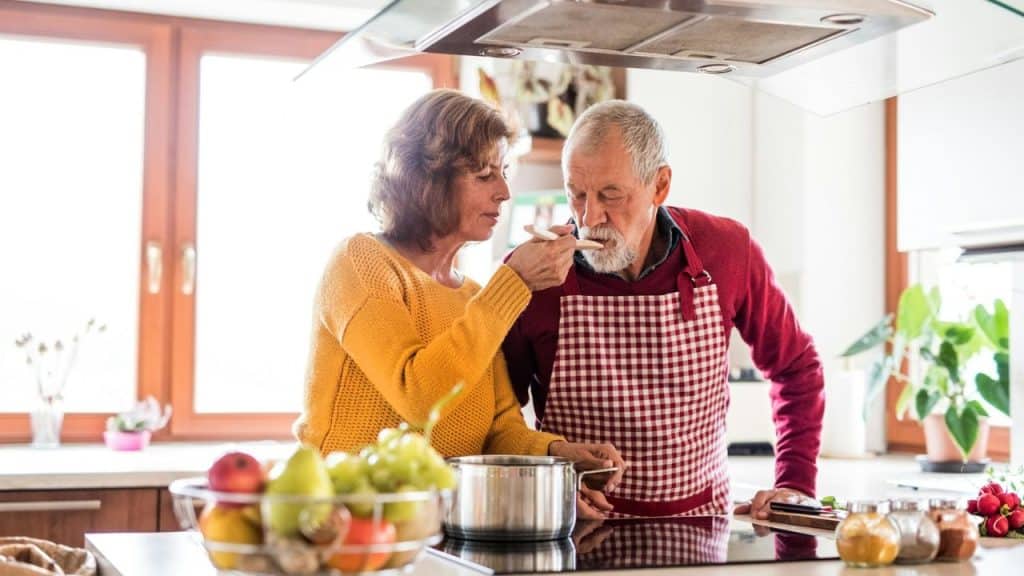 A woman in a yellow sweater feeds a spoonful of food to a man in a red apron.