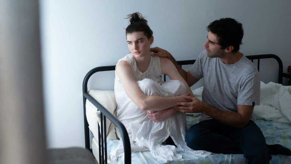 A man sits on a bed and places his hand on the shoulder of a woman.
