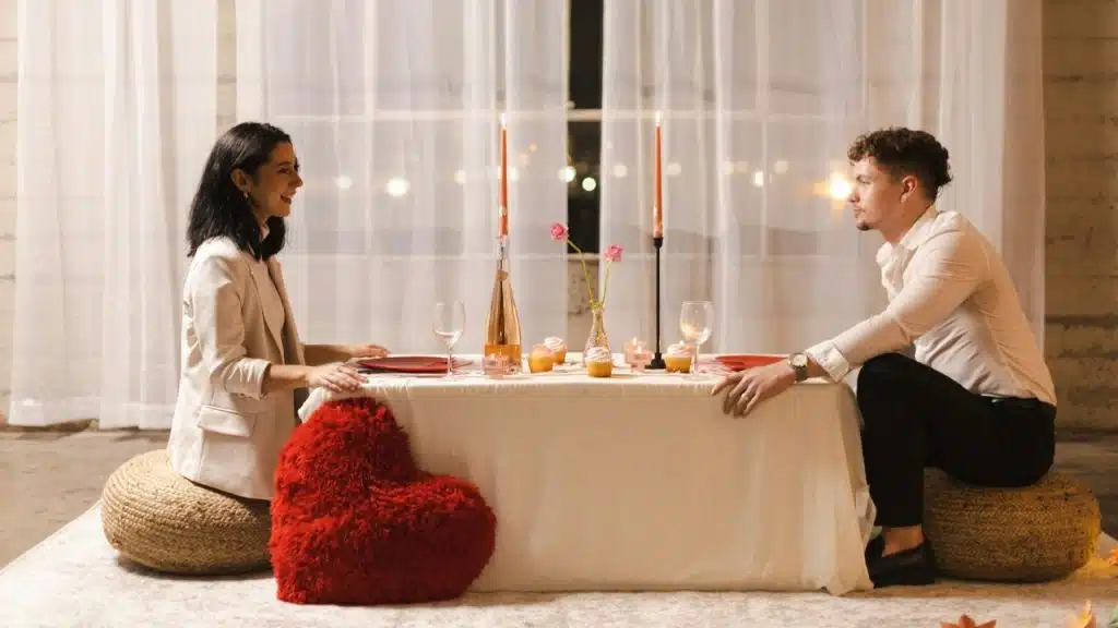 A man and woman sit on floor cushions facing each other at a candlelit table.