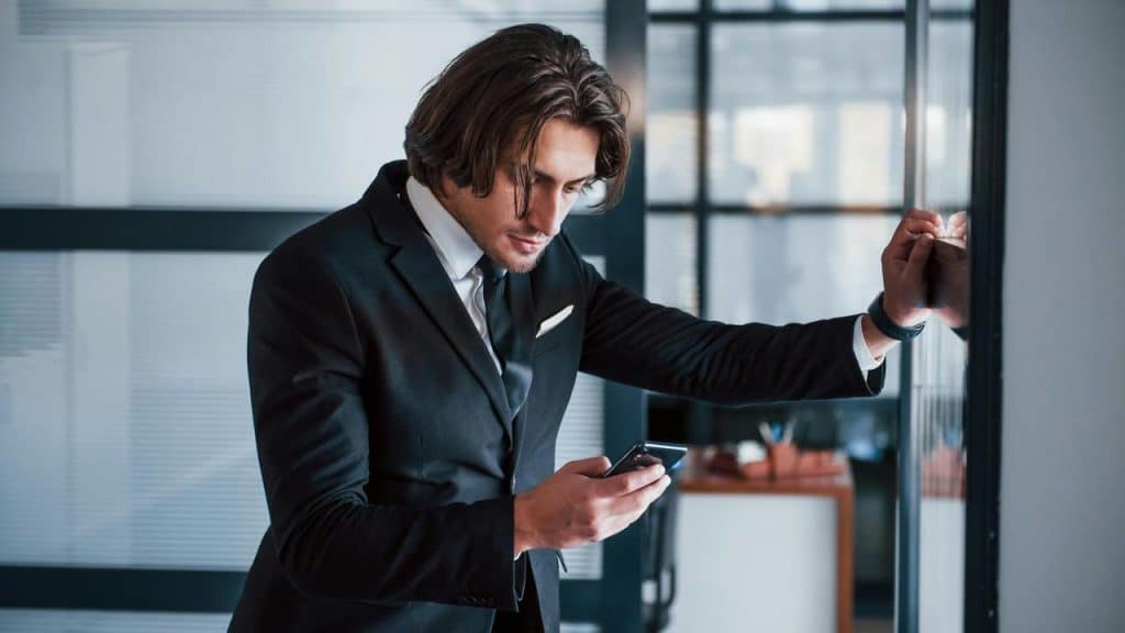 A businessman in a suit checking his phone while leaning against a glass wall.