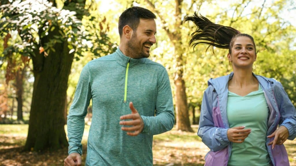 A man and woman in athletic wear run together through a sunlit, wooded park.