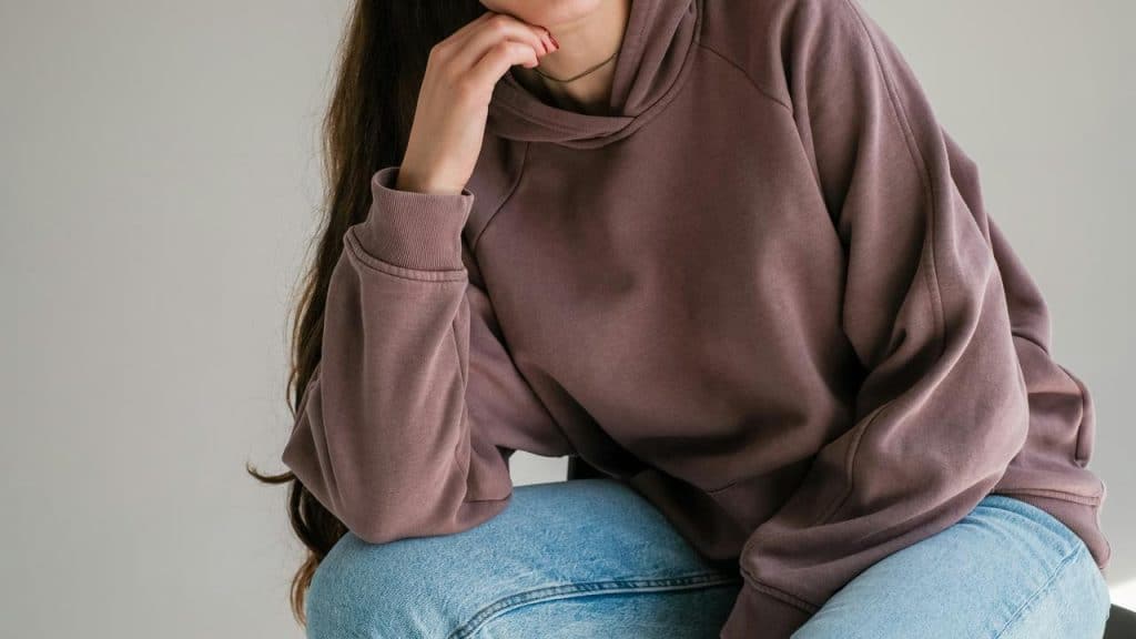 A person wearing a loose hoodie and jeans, seated with a hand near their chin.