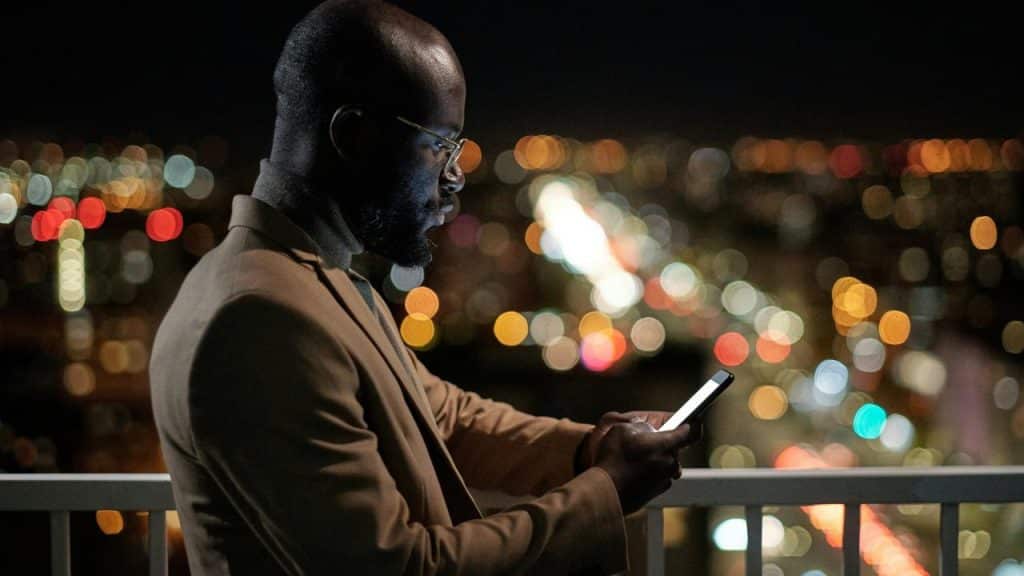 A man using a smartphone on a balcony overlooking city lights at night.