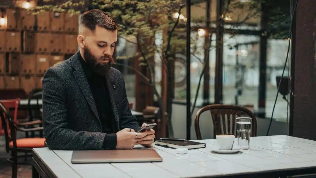 A man in a suit sits at a café table using his phone with a laptop.