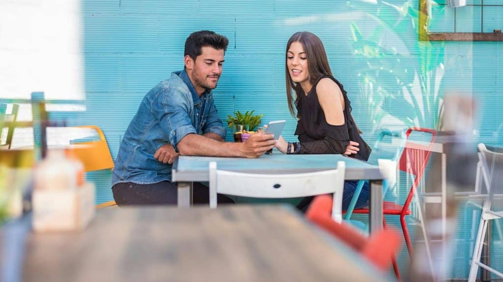 A man and woman sit at a café table smiling while looking at a phone together.
