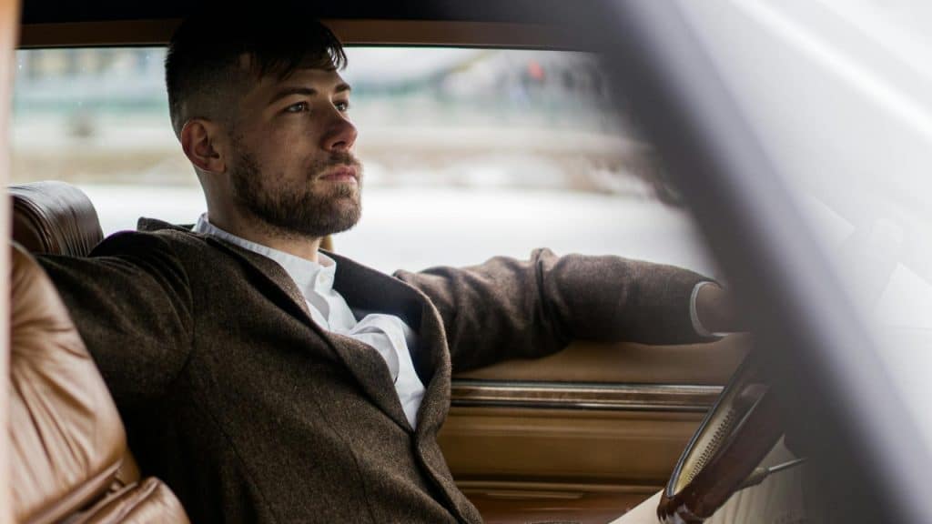 A man with a beard drives a car while looking ahead through the front windshield.
