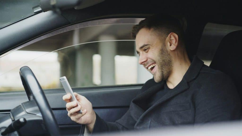 A man sitting in the driver's seat of a car laughs while looking at his phone.