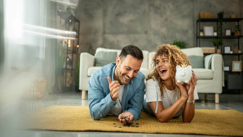 A laughing man and woman lie on a rug with a piggy bank and coins.