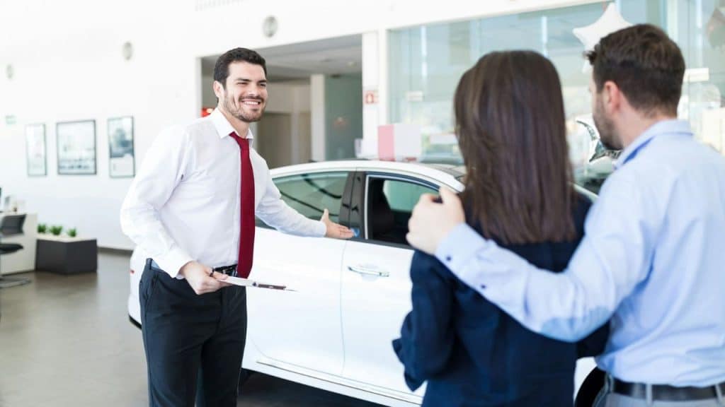 A smiling salesman in a red tie gestures toward a white car for a couple.
