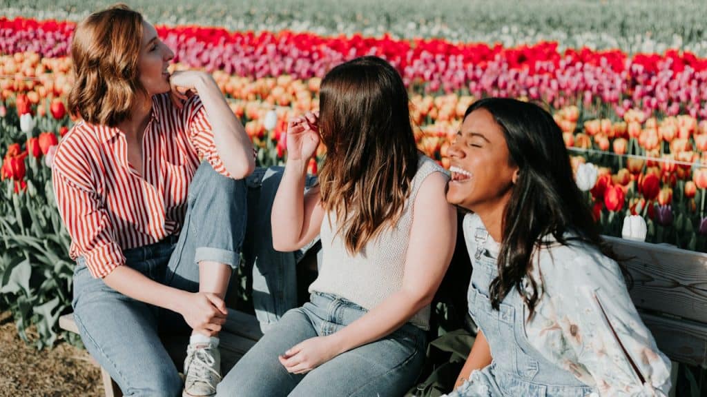 A trio of friends sitting on a bench in front of a flower field and talking while one of them laughs.
