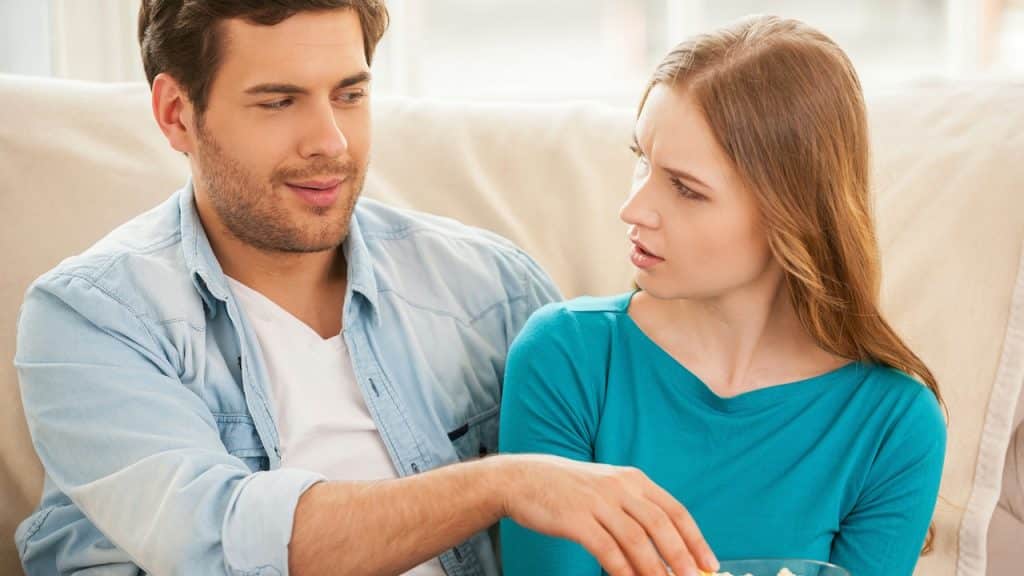 Young couple on a couch, man holding popcorn, woman looking at him with a frown or skeptical expression.